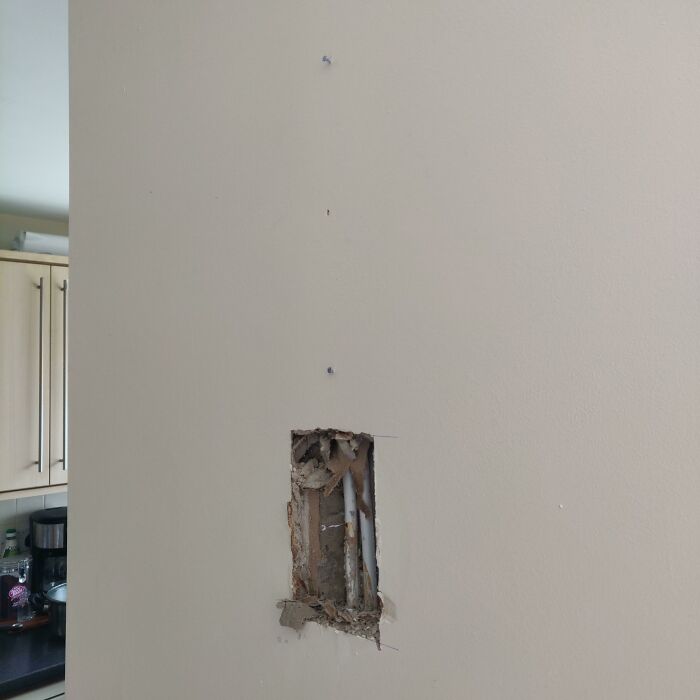 Wall with a hole exposing pipes, possibly left by in-laws; kitchen background visible.