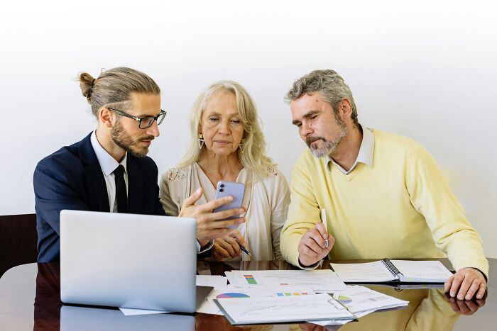 Three colleagues discuss office expressions overused in meetings, reviewing paperwork and a smartphone together.