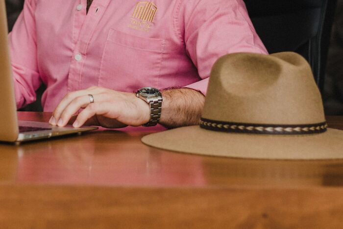 Man in pink shirt using laptop with a watch on wrist, brown hat on desk. Office setting, focus on overused expressions.