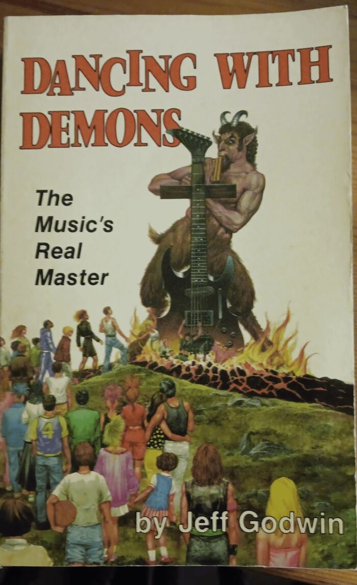 Terrible book cover of "Dancing with Demons" by Jeff Godwin, featuring a demon playing a guitar amidst fire and people looking on.
