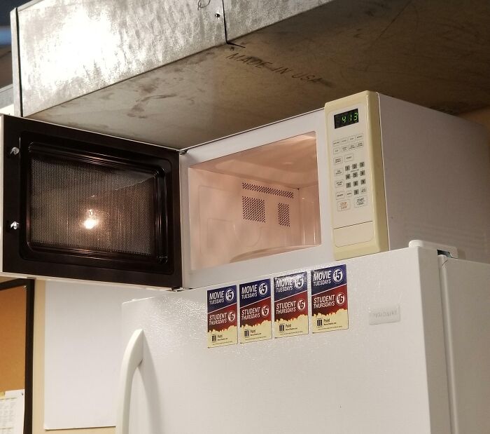 Microwave with open door placed awkwardly on top of a fridge, showing a common office scenario with challenging colleagues.