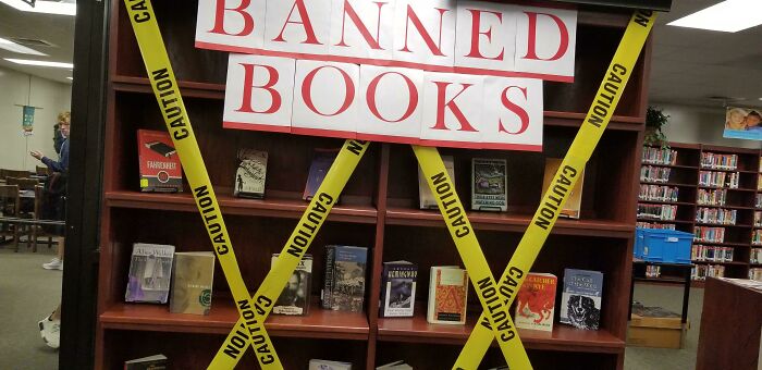Library shelf with "Banned Books" sign and caution tape, highlighting issues in American schools.