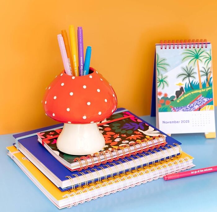 Mushroom pen holder atop colorful notebooks, next to a vibrant desk calendar.
