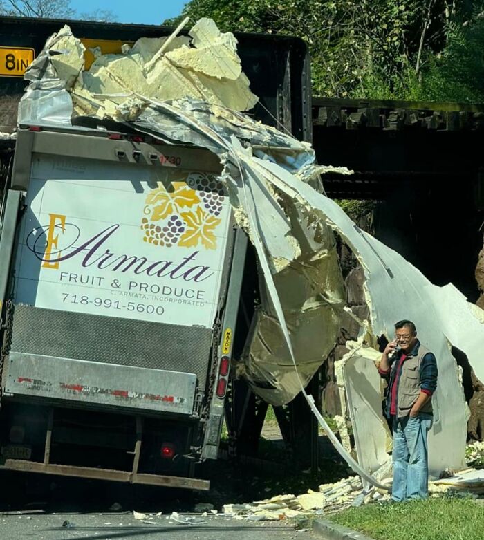 Truck stuck under bridge, severely damaged; driver on phone looking stressed, epitomizing worst day at work.