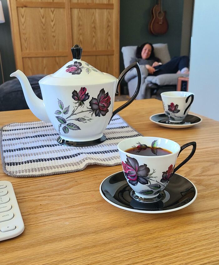 Vintage floral teapot and cups on table; person relaxing on sofa, guitar on wall in the background.