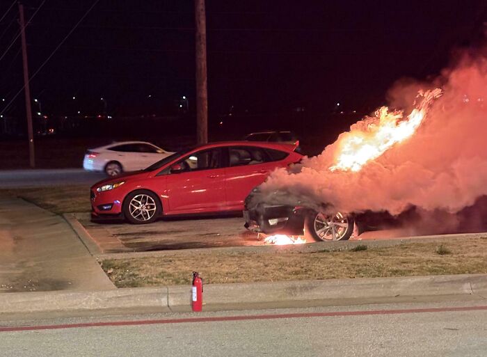 Car on fire in parking lot at night, signifying worst day at work. Nearby red car, fire extinguisher on ground.