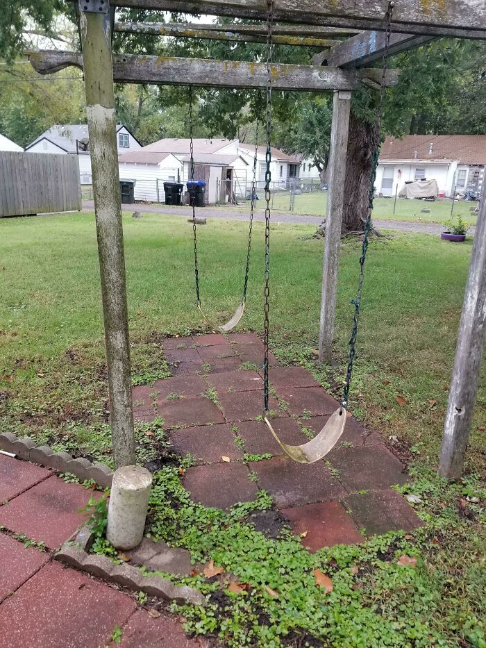 Broken swings on a neglected playground, showcasing disastrous design with uneven pavers and overgrowth.