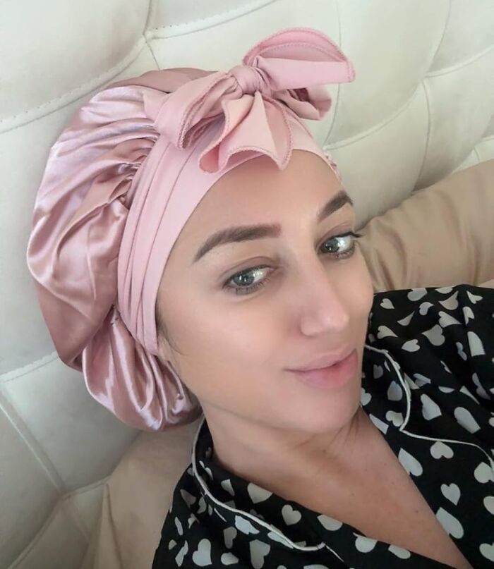 Woman with curls wearing a pink satin hair wrap, smiling and relaxing, enhancing curly hair care routine.
