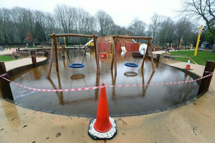 Flooded playground design error with swings submerged, surrounded by caution tape and traffic cone.