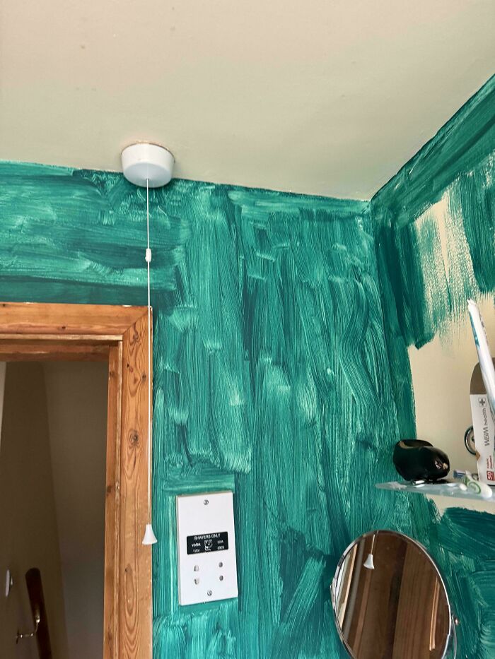 Poorly painted green wall with a pull cord, a white switch, and a mirror reflecting a shelf.