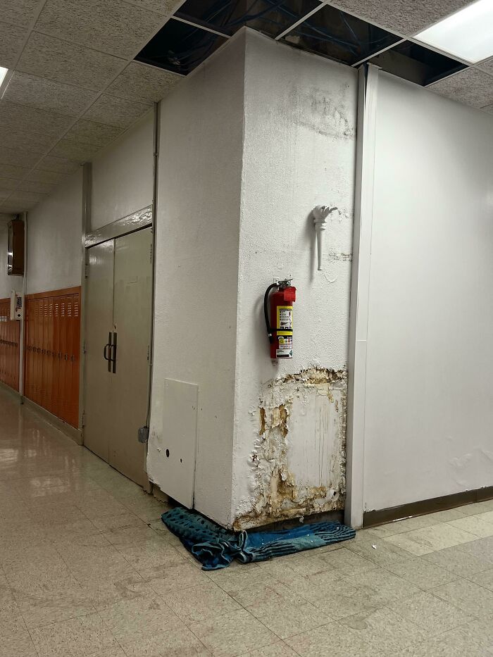 Damaged wall corner in an American school hallway, showing peeling paint and exposed material near lockers.