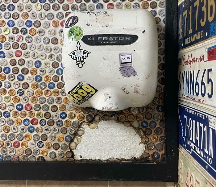 Hand dryer on a wall of bottle caps with worn-down plaster beneath, showing time's effect on belongings.
