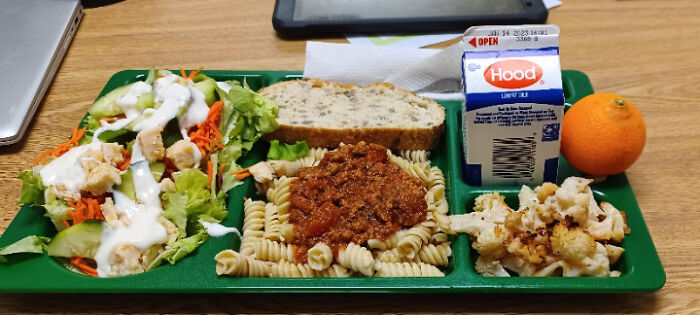 School lunch tray with pasta, salad, bread, milk carton, and orange, showcasing American school meal variety.