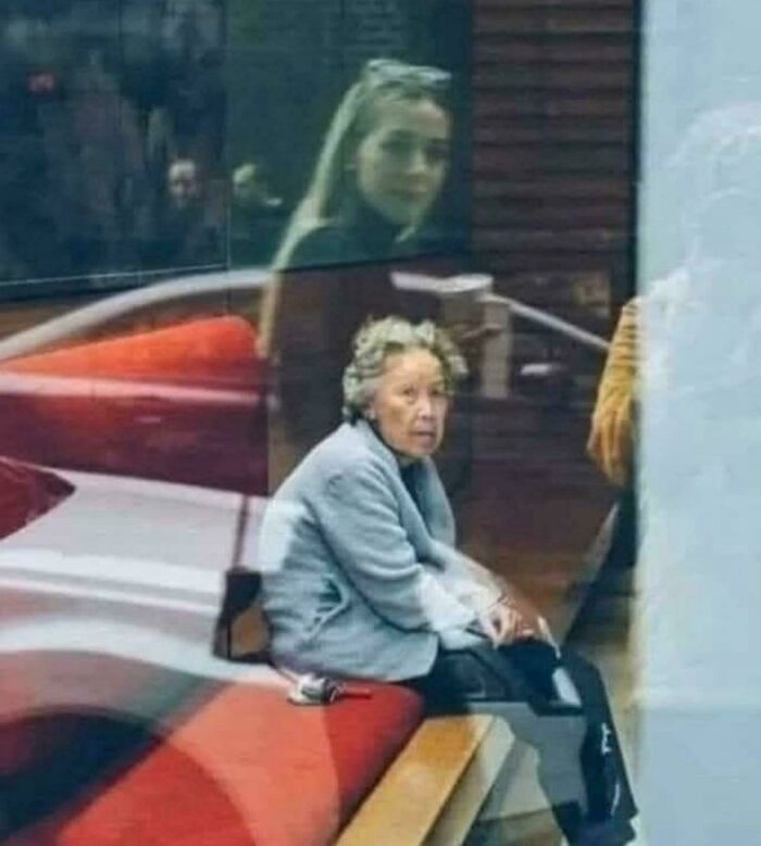Elderly woman sitting on a bench indoors, with reflections in the glass; a seemingly uninteresting moment captured.
