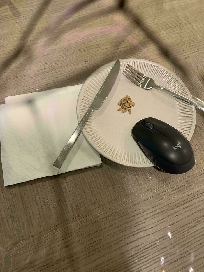 Plain plate with a few seeds, knife, fork, napkin, and a computer mouse on a wooden table.