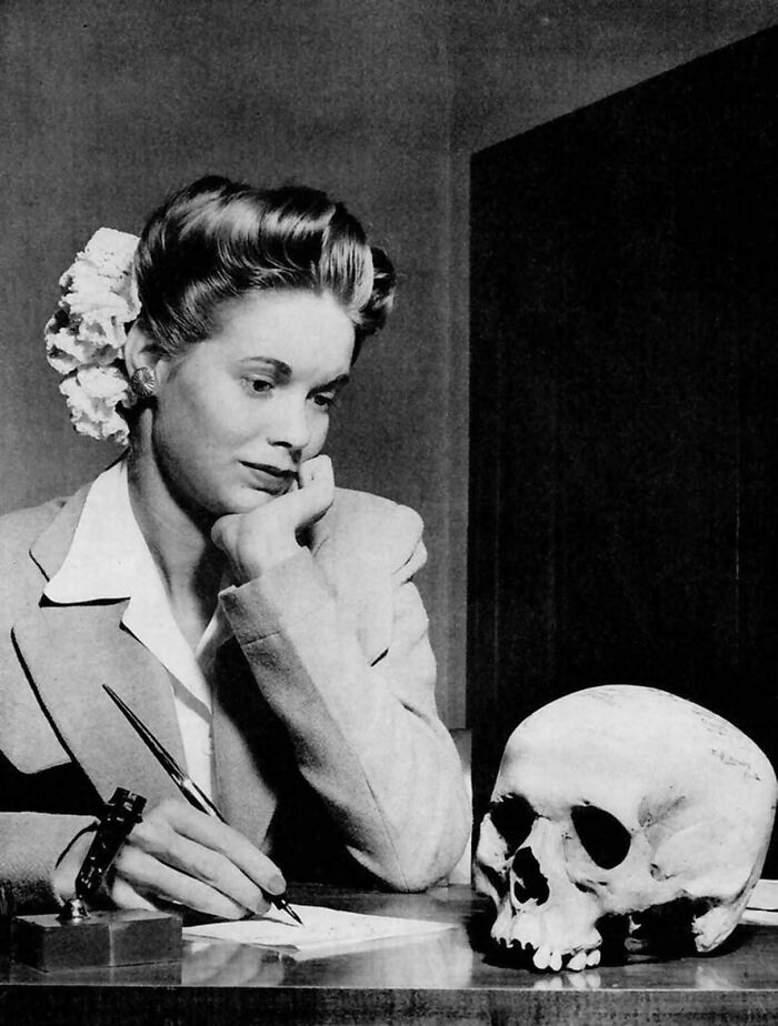A woman in vintage attire pensively writes beside a human skull, embodying unsettling past imagery.
