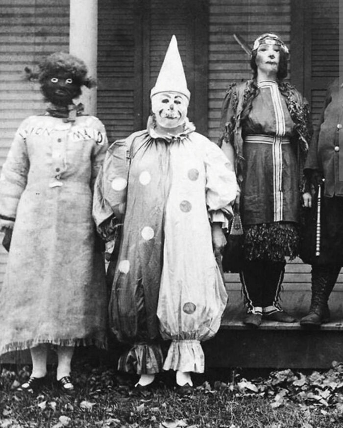 Black and white photo of unsettling clowns in vintage costumes standing on a porch.