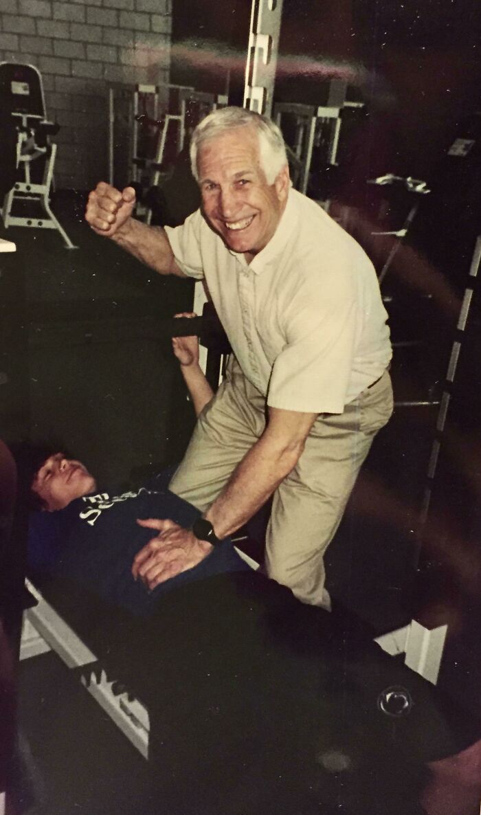 Elderly man in gym lifting hand weights, smiling, with a young man assisting, in a slightly unsettling photo from the past.