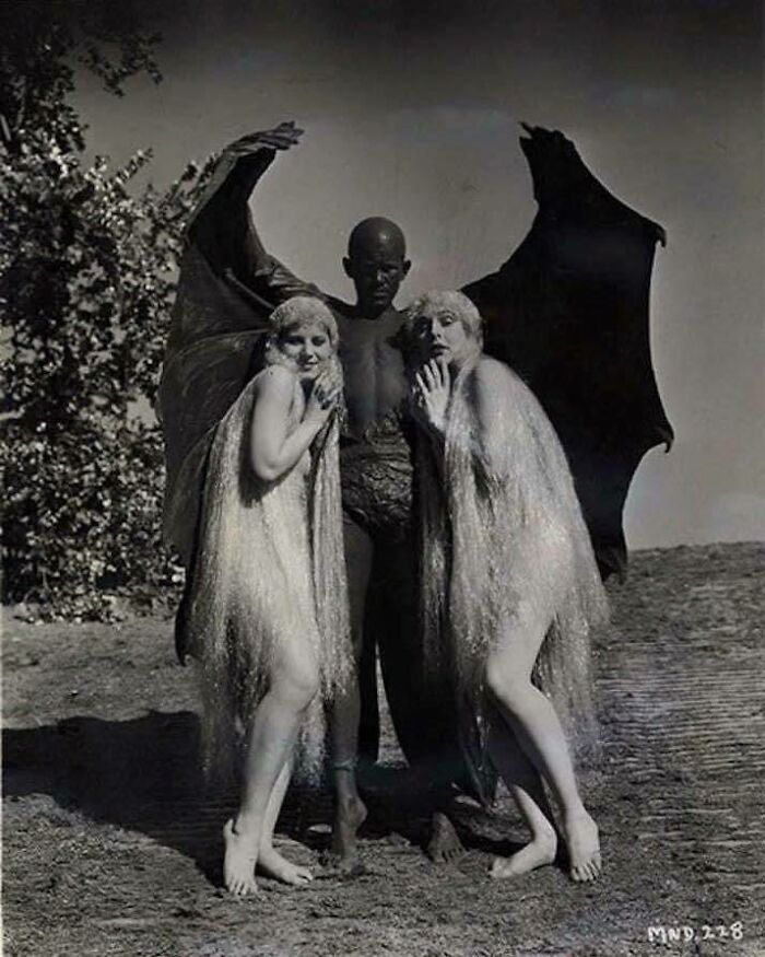 Vintage photo of two women in ethereal dresses, posing with a winged figure, capturing an unsettling scene from the past.
