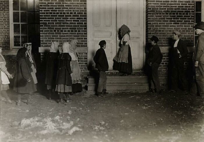 Vintage photo of people in period clothing standing outside a brick building, capturing an unsettling moment from the past.