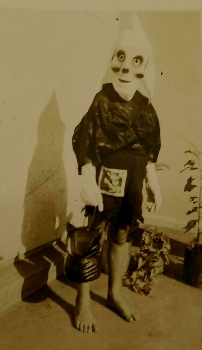 A person in an unsettling, vintage ghost costume, holding a mask, stands barefoot in a corner, casting a shadow.