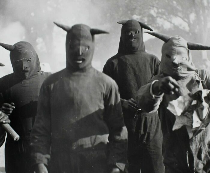 Unsettling historical photo shows masked figures in dark costumes outdoors, exuding a mysterious aura.