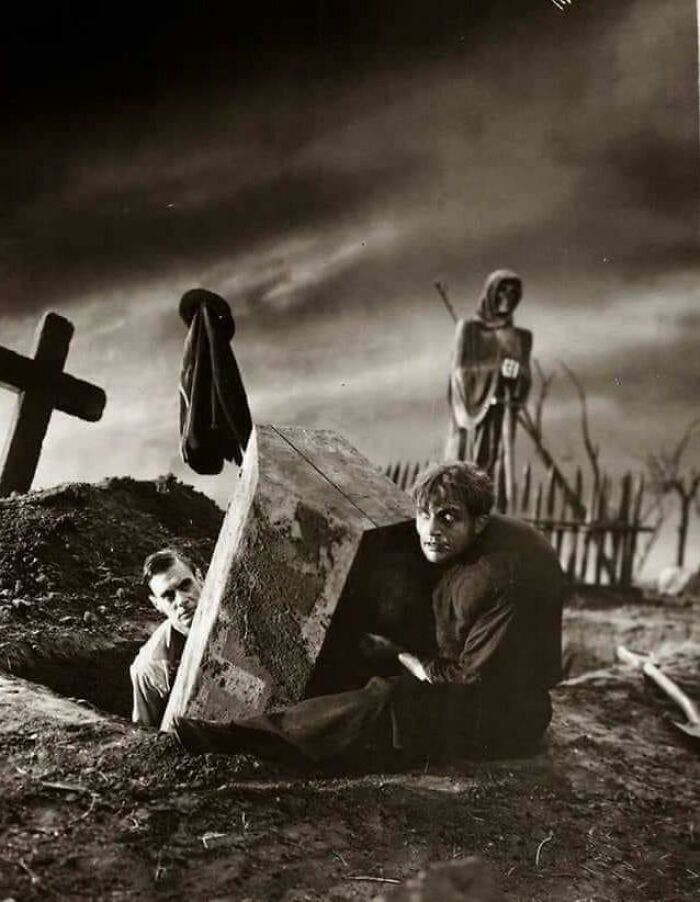 Two men in a graveyard scene, one emerging from a coffin, surrounded by unsettling old grave markers and a scarecrow.