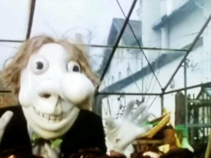 A large puppet with a wide grin in a greenhouse setting, part of unsettling historical photos collection.