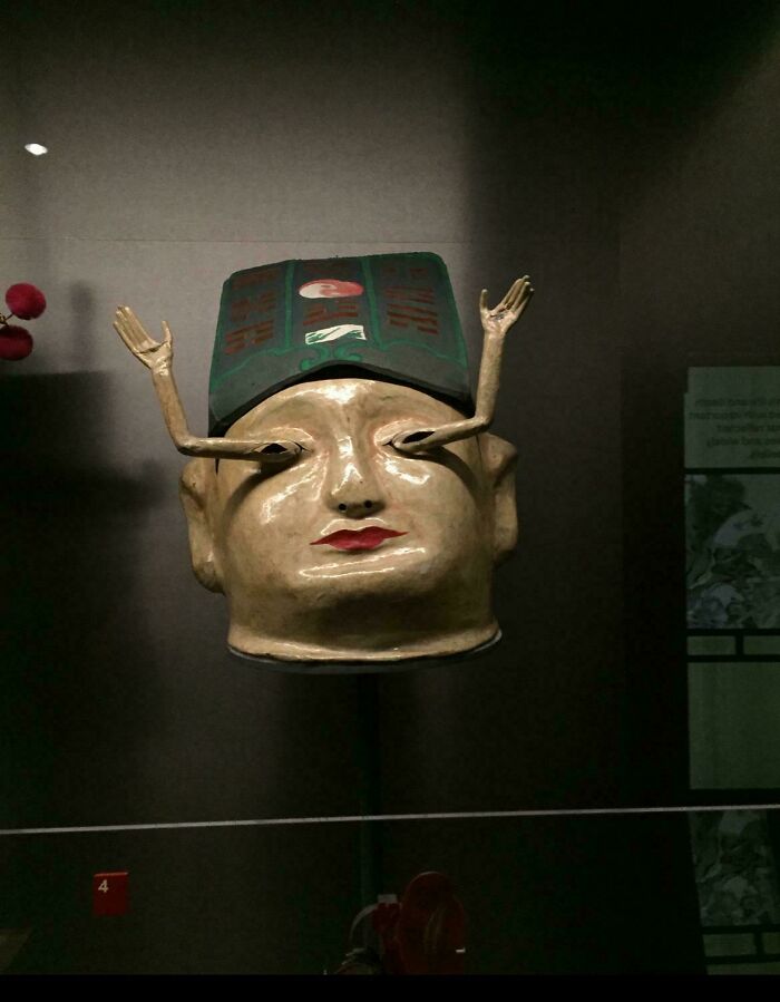 Unsettling sculpture of a head with arms in a museum display, featuring vivid colors and exaggerated features.