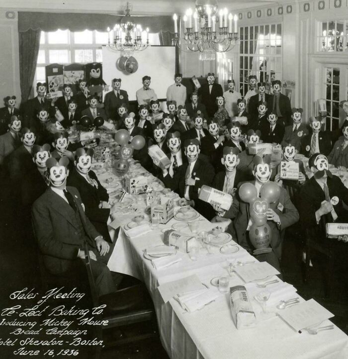 Unsettling vintage photo with people in Mickey Mouse masks at a corporate meeting, 1936 pastries and balloons visible.