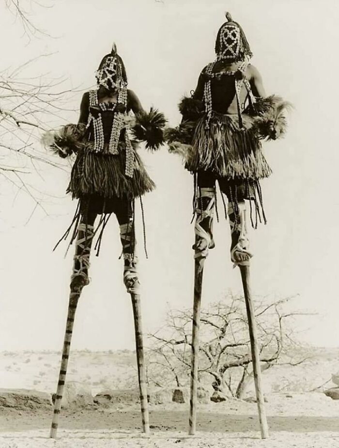 Two people in tribal costumes on stilts, creating an unsettling historical scene in a barren landscape.
