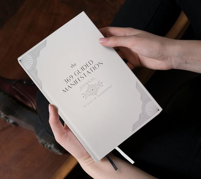 Person holding a white manifestation journal called "369 Guided Manifestation" with decorative design.