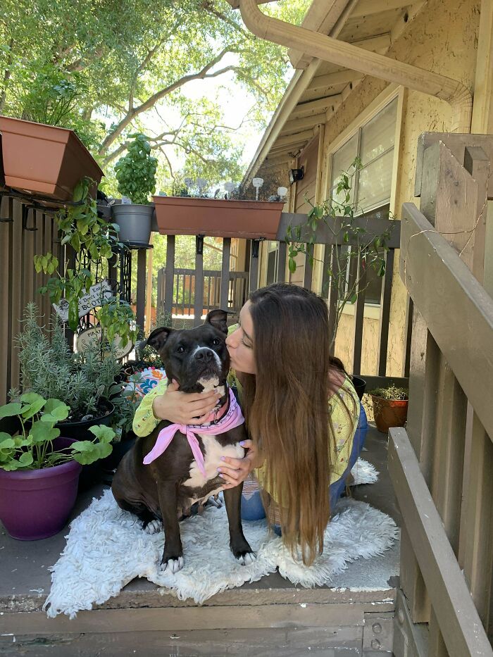 Woman hugs and kisses her rescued dog on a porch, surrounded by potted plants.