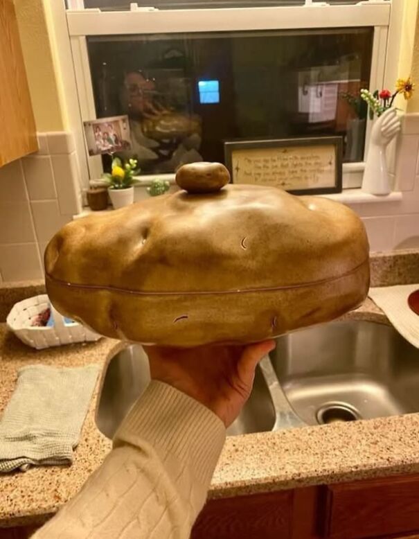 Person holding a large, unusual pottery item resembling a hat, with a kitchen sink and window in the background.