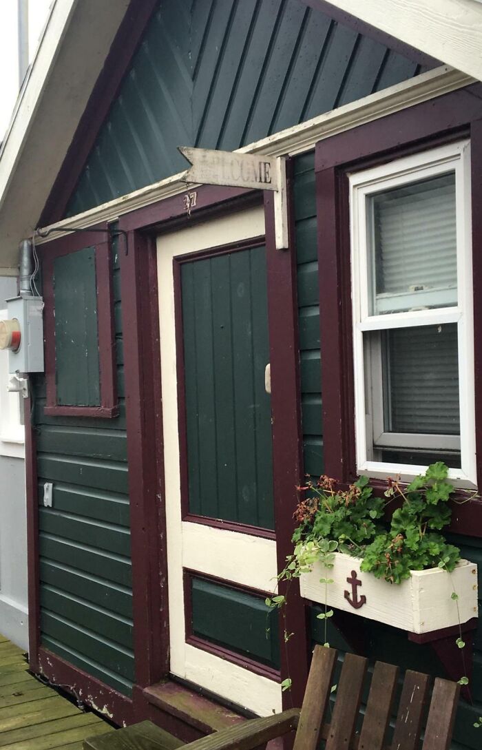 Worn-down house facade with green paneling, faded "Welcome" sign, and weathered plants in a window box.
