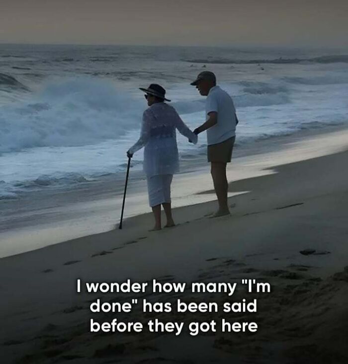 Elderly couple on beach holding hands, embracing a 'sadcastic' moment by the waves with a cane.