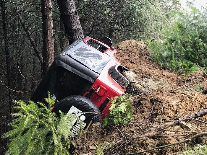 Overturned red Jeep in a forest area, resting against trees and covered in mud.