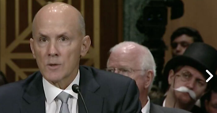 Bald man speaking at a formal event, while a person in a top hat and fake mustache watches from the background.