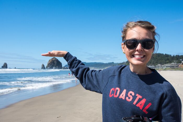 Smiling woman at a stunning beach, wearing sunglasses and a "Coastal" sweatshirt, with ocean waves and rock formations in view.