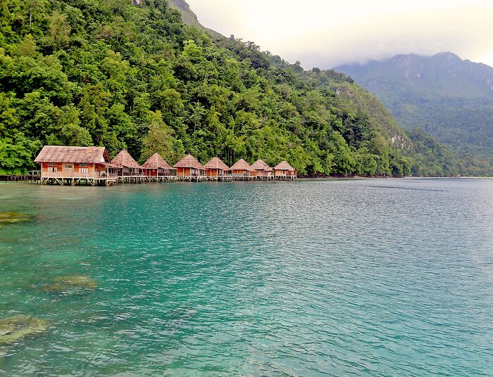 Huts over crystal clear water on a stunning beach, surrounded by lush greenery and mountains.