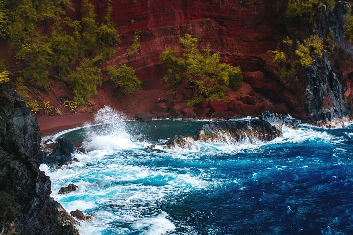 Stunning beach with vibrant blue waves crashing against a rocky shore under a rugged, red cliff.