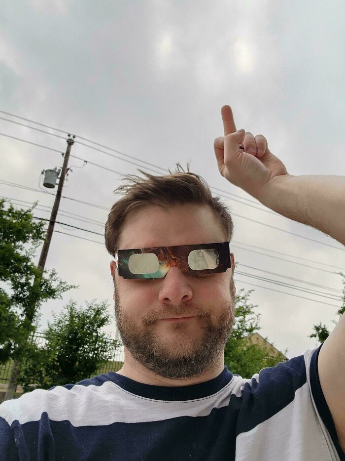 Man wearing eclipse glasses under cloudy sky, pointing upwards, having a humorous day.