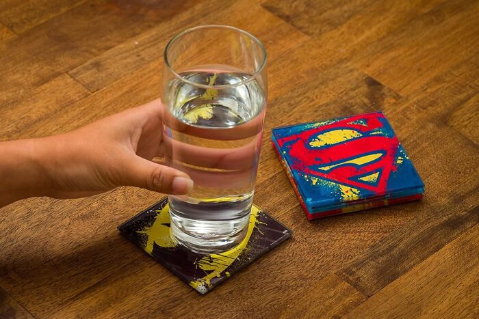 Hand holding a glass of water on a superhero-themed coaster on a wooden table, showcasing Amazon store fandom design.