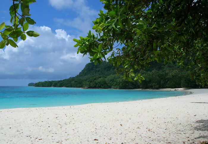 Tropical beach with white sand and turquoise water, lush greenery in the background, under a cloudy sky.