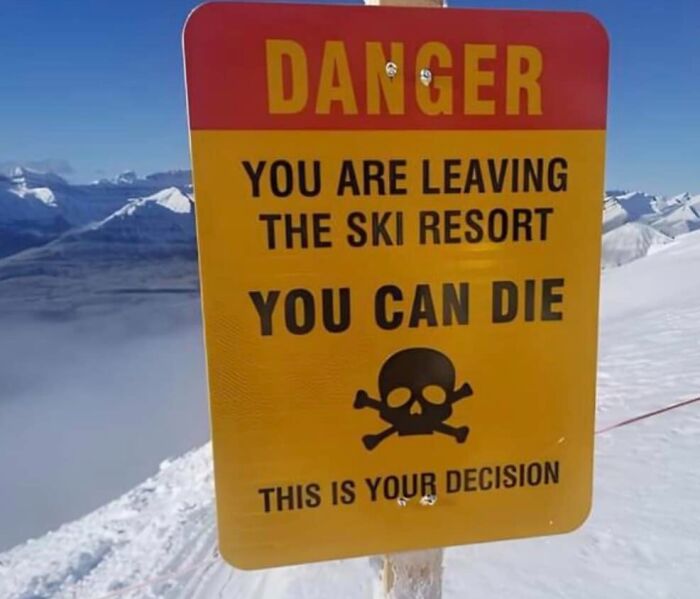 Warning sign at ski resort exit, cautioning potential danger with skull icon, snowy mountain backdrop.