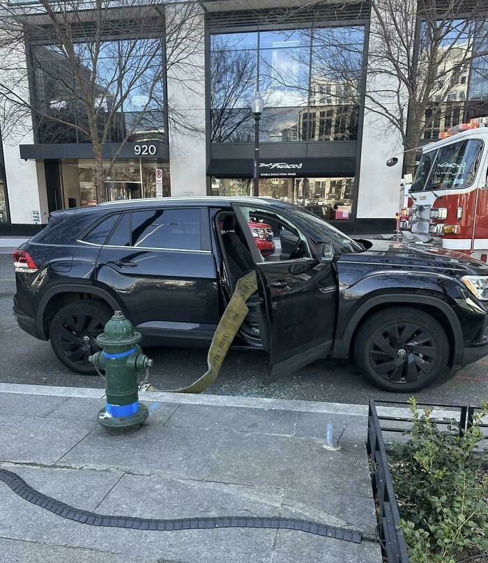Coche negro estacionado junto a hidrante con manguera de bomberos atrapada en la puerta, personas con mal día.