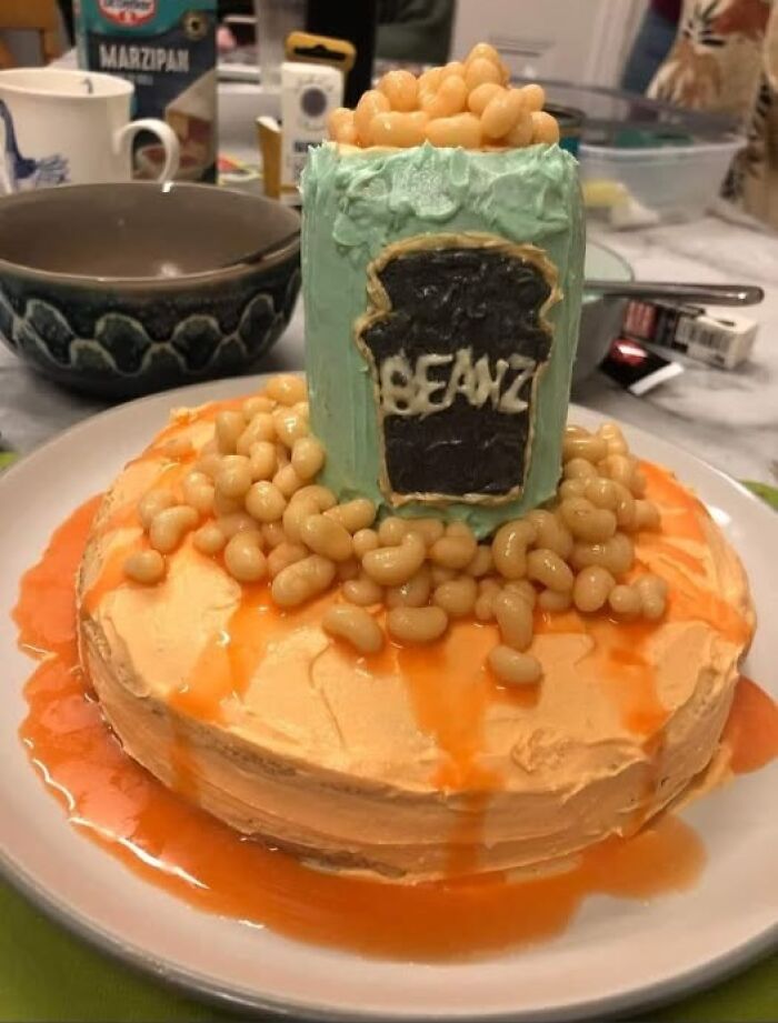 Cake resembling a can of beans, with icing and spilling baked beans on top, showcasing a funny baking fail.