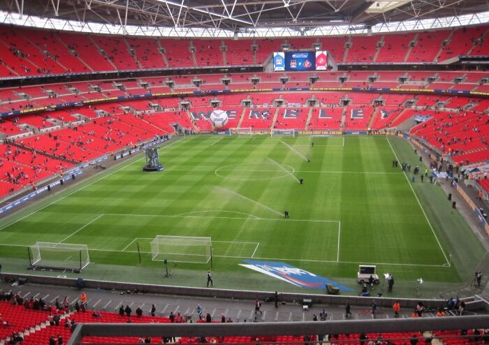 A wide view of a famous soccer stadium with green pitch and red seats, one of the cathedrals of soccer legends.