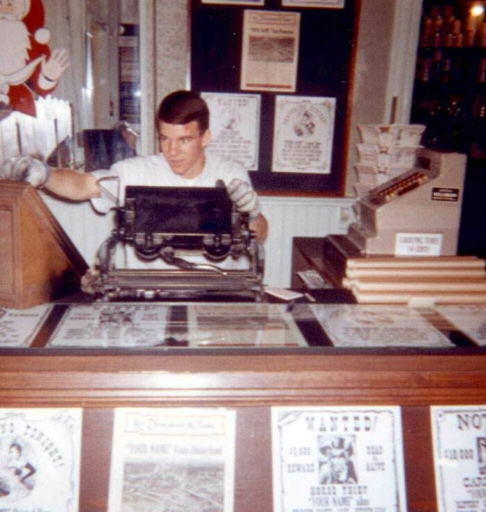 Man working a printing press in a retro American shop during the JFK era, surrounded by vintage prints and posters.