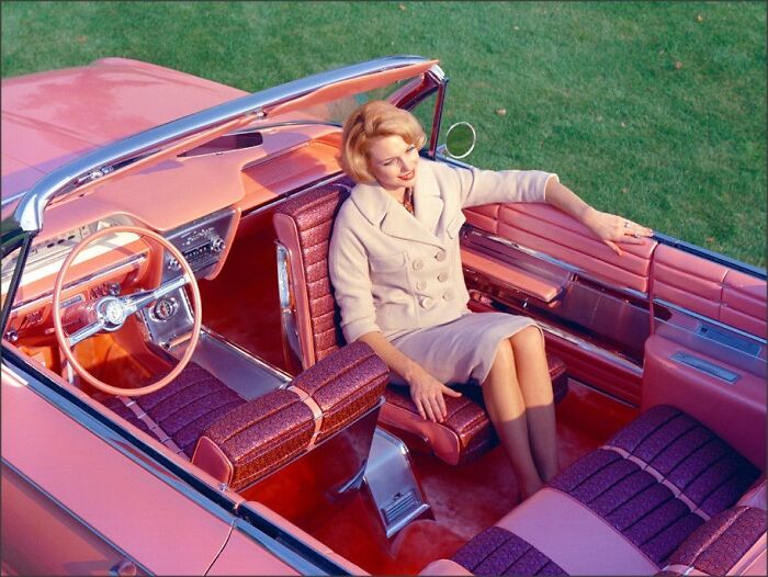 Woman in vintage pink convertible car, wearing a beige coat, representing America during the JFK years.